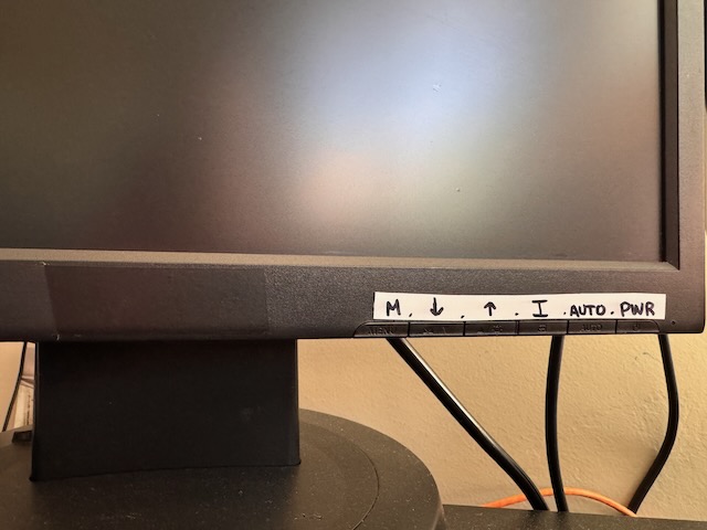 A close up photo of a monitor with black electrical tape covering the brand logo and white electrical tape with sharpie written on it indicating functions of unlabelled buttons.